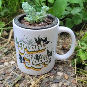 May include: White ceramic mug with the text "Plant Lady" in a retro font, filled with soil and a small succulent plant. The mug has a black rim and a curved handle. The background is a mix of green foliage and small stones.