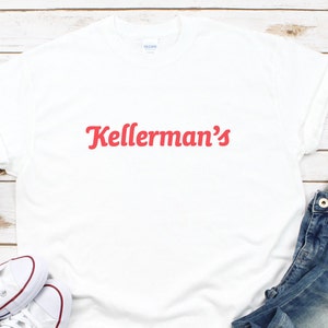 Op de afbeelding: Wit t-shirt met de rode tekst "Kellerman's" op de voorkant. Het shirt is opgevouwen, met een paar witte sneakers met rode rand en een blauwe jeans op de achtergrond. Het shirt is gemaakt van een zacht materiaal.