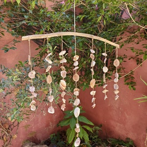 Peut inclure: Mobile artisanal de coquillages suspendu &agrave; une branche de bois incurv&eacute;e. Le mobile est compos&eacute; de coquillages de diff&eacute;rentes tailles et formes, reli&eacute;s par de la ficelle naturelle. Les coquillages sont principalement blancs et beiges, pour un style c&ocirc;tier.