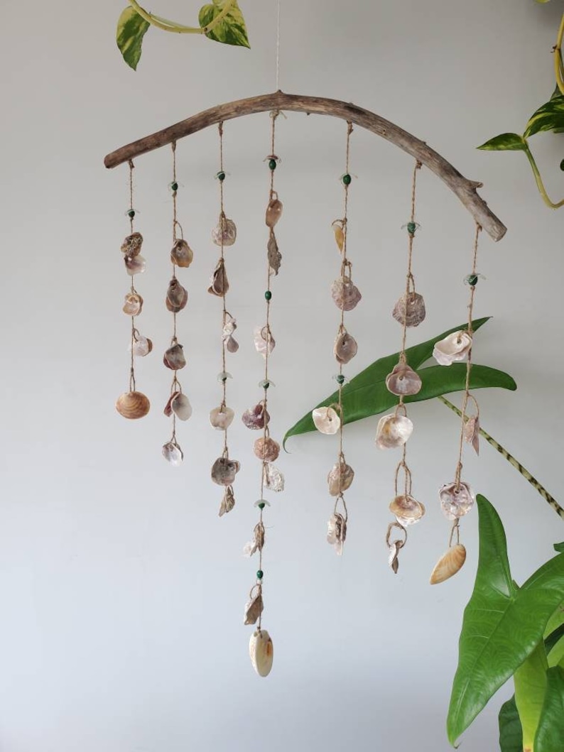 Peut inclure: Mobile d&eacute;coratif fait &agrave; la main avec des coquillages. Une branche de bois incurv&eacute;e suspend plusieurs brins de coquillages et de petites perles vertes. Les coquillages varient en taille et en couleur, allant du cr&egrave;me au brun clair. Le mobile est suspendu devant un mur blanc avec des plantes vertes.