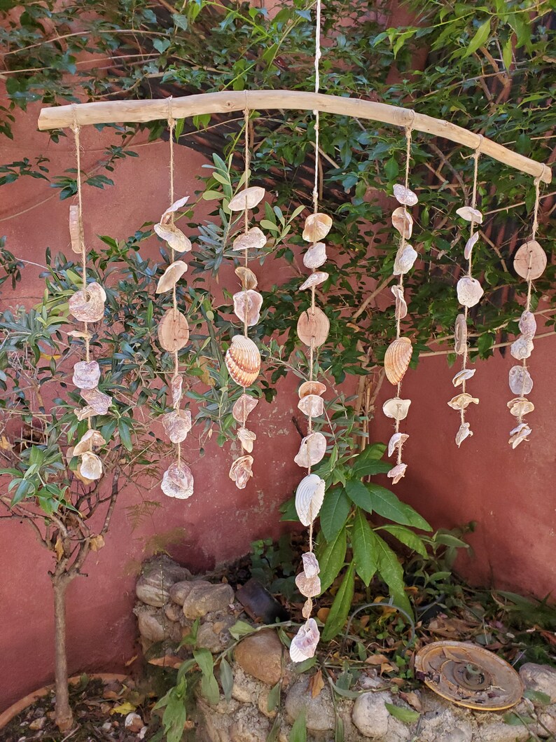 Peut inclure: Un carillon &eacute;olien artisanal en coquillages. Le carillon est compos&eacute; de divers coquillages et disques en bois suspendus &agrave; une branche en bois naturel avec de la ficelle. Les coquillages sont dans des tons de blanc, beige et marron.