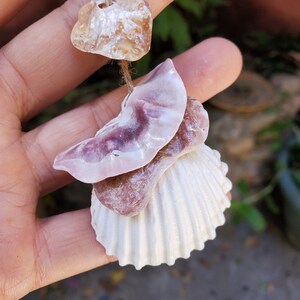 Peut inclure: Mobile artisanal de coquillages et de pierres naturelles. Il comprend une coquille Saint-Jacques blanche, une coquille rose-violet et plusieurs petites pierres brun clair. Les coquillages et les pierres sont enfil&eacute;s sur de la ficelle naturelle.