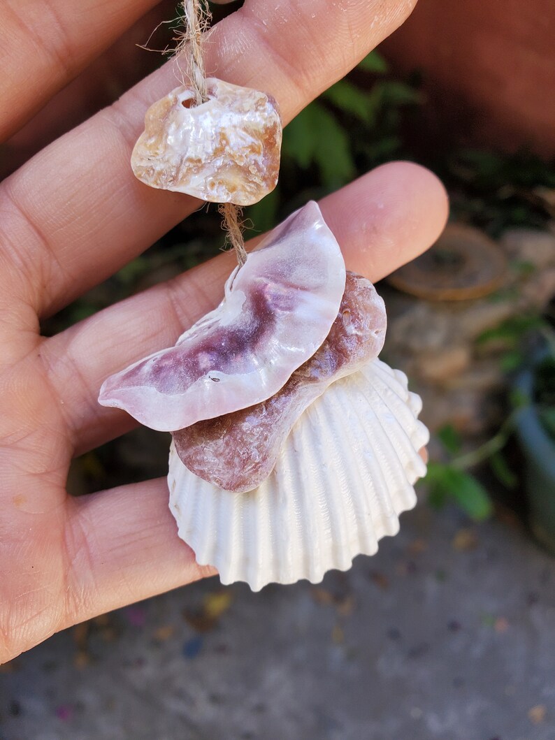 Peut inclure: Mobile artisanal de coquillages et de pierres naturelles. Il comprend une coquille Saint-Jacques blanche, une coquille rose-violet et plusieurs petites pierres brun clair. Les coquillages et les pierres sont enfil&eacute;s sur de la ficelle naturelle.