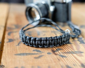 May include: A black and grey braided camera strap with a silver metal clasp.