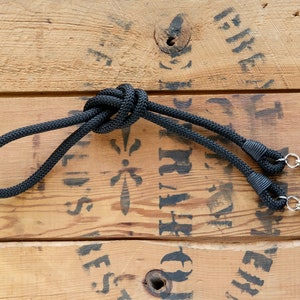 May include: Black camera strap with a knotted design and two metal clips. The strap is lying on a wooden surface.