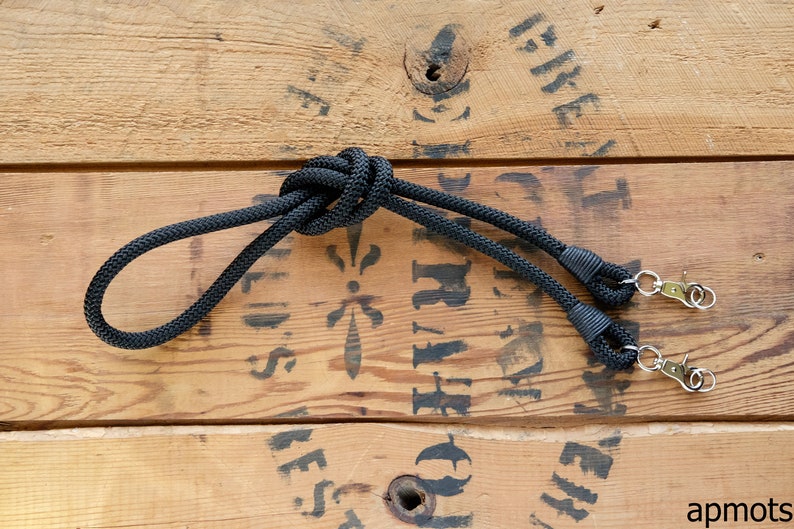 May include: Black camera strap with a knotted design and two metal clips. The strap is lying on a wooden surface.