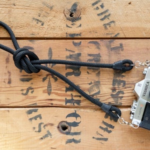 May include: A black camera strap with a knot and metal clasps attached to a silver camera with a black lens. The camera is labelled "YASHICA" and "Electro 35 GS". The camera and strap are on a wooden surface.