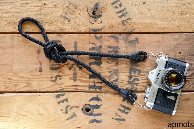 May include: A black camera strap with a knot and metal clasps attached to a silver camera with a black lens. The camera is labelled "YASHICA" and "Electro 35 GS". The camera and strap are on a wooden surface.
