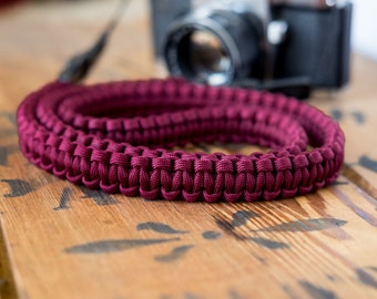 May include: A close-up of a maroon camera strap made of braided paracord. The strap is coiled and lying on a wooden surface.