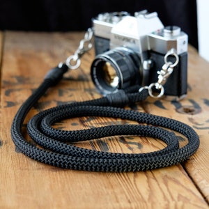 May include: A black camera strap made of thick, braided rope with silver metal clasps. The strap is lying on a wooden surface.