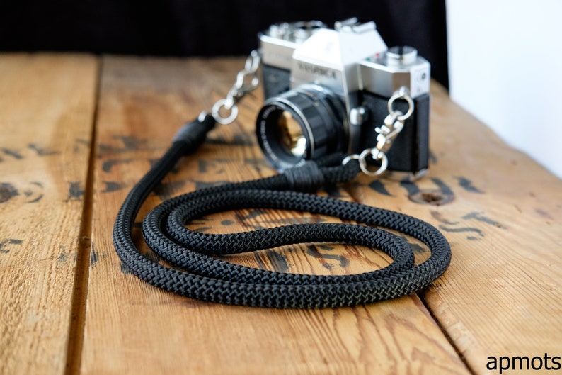 May include: A black camera strap made of thick, braided rope with silver metal clasps. The strap is lying on a wooden surface.