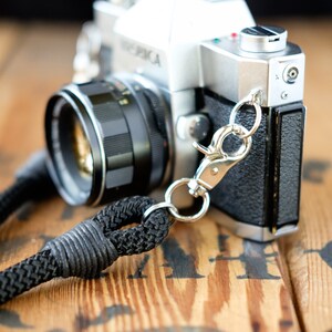 May include: A silver and black 35mm film camera with a black leather strap attached to a silver metal clip. The camera is sitting on a wooden surface.