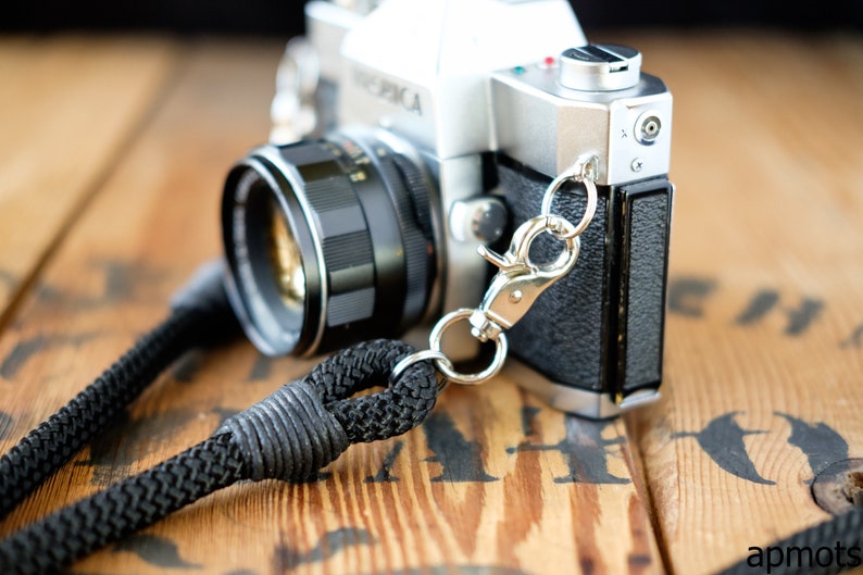 May include: A silver and black 35mm film camera with a black leather strap attached to a silver metal clip. The camera is sitting on a wooden surface.