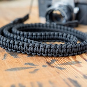 Op de afbeelding: Een zwarte cameraband gemaakt van gevlochten paracord. De band is opgerold en ligt op een houten oppervlak.