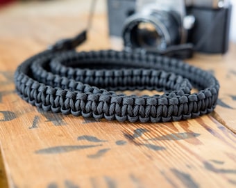 May include: A black camera strap made of braided paracord. The strap is coiled and lying on a wooden surface.