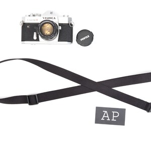 May include: A black camera strap with silver metal clasps. The strap is crossed and laid flat on a white surface. A black and white camera and a black lens cap are visible in the background.