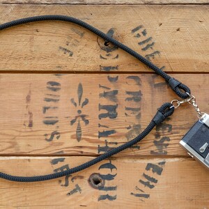 May include: A black rope camera strap with a silver camera attached to it. The camera is a Yashica Electro 35 GS. The strap and camera are on a wooden surface.