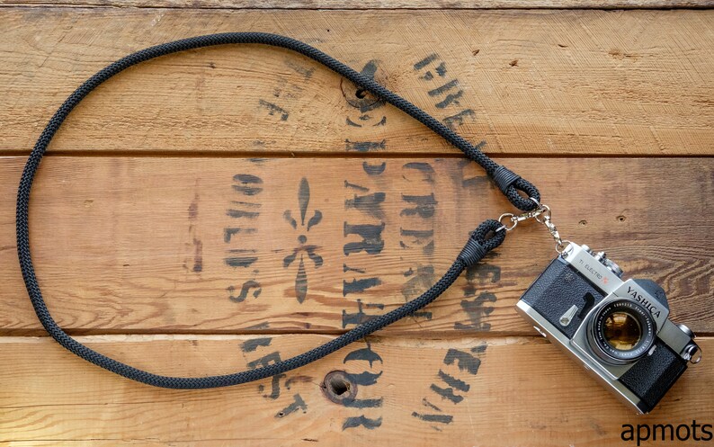 May include: A black rope camera strap with a silver camera attached to it. The camera is a Yashica Electro 35 GS. The strap and camera are on a wooden surface.