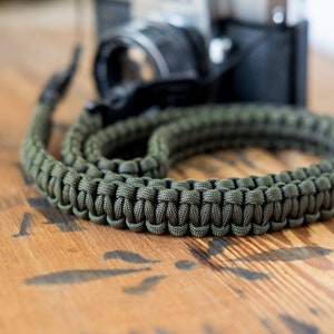 May include: A green paracord camera strap, woven in a tight pattern, lies on a wooden surface. The strap is designed for a camera and is made of durable paracord.