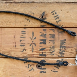 May include: A black camera strap made of braided rope with metal clasps. The strap is hanging from a silver camera with a black lens on a wooden surface.