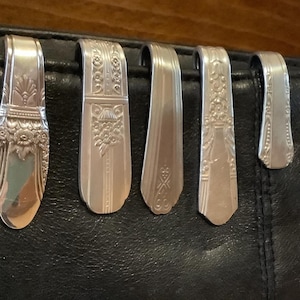 May include: A collection of silver spoon handles, repurposed as bag clips, are displayed against a black leather surface. Each clip features a unique embossed design, showcasing floral and geometric patterns. The spoons are bent to form a hook shape.
