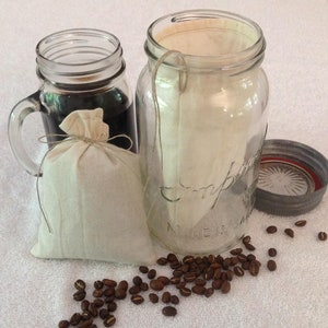 May include: Two glass jars with lids, one filled with coffee grounds in a white muslin bag, the other with a white muslin filter bag. Coffee beans are scattered on a white surface.