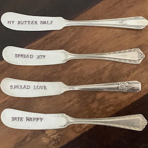 May include: Four silver butter spreaders with stamped text. The text on the spreaders reads: "MY BUTTER HALF", "SPREAD JOY", "SPREAD LOVE", and "BRIE HAPPY". The spreaders have decorative handles and are arranged on a wooden surface.