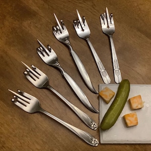 May include: A collection of stainless steel forks with a unique design, arranged on a wooden surface. The forks are paired with a pickle and cheese cubes on a white square plate. The forks' design resembles a hand gesture.