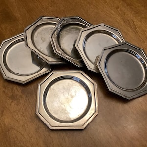 May include: Six silver-coloured, octagonal plates are arranged on a wooden surface. Each plate has a raised rim and a smooth, reflective surface. The plates' geometric design and metallic sheen are highlighted. These plates are likely for serving food or decorative purposes.