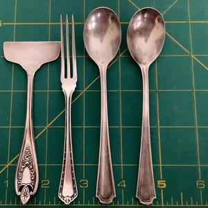 May include: A collection of antique silverware, including a serving spatula, a fork, and two spoons. The utensils are made of silver-colored metal and rest on a green cutting mat. The spatula has an ornate handle design.