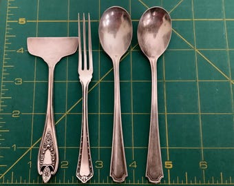 May include: A collection of antique silverware, including a serving spatula, a fork, and two spoons. The utensils are made of silver-colored metal and rest on a green cutting mat. The spatula has an ornate handle design.