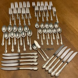 May include: A comprehensive set of vintage silverware, comprising forks, spoons, and knives, displayed on a wooden surface. The cutlery features decorative handles and a polished finish, suitable for formal dining. The set includes various serving utensils.