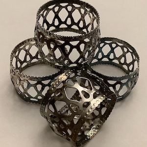 May include: Four silver metal napkin rings with an openwork design. The rings are arranged in a circular pattern, with one ring partially overlapping the others. The metal has a textured appearance, and the design features a series of interconnected shapes.