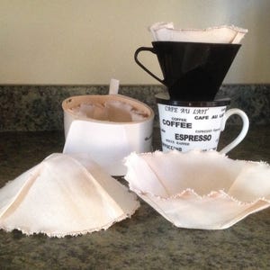 May include: Three white cotton coffee filters, two are cone shaped and one is a flat disc. The filters are sitting on a counter next to a white coffee mug with the text "Cafe au Lait, Coffee, Espresso" printed on it.