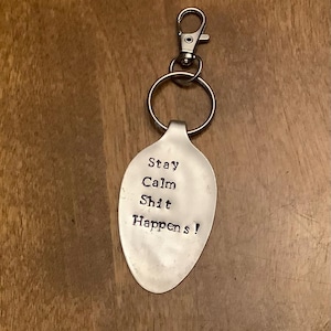 May include: A silver spoon keychain with the text "Stay Calm Shit Happens!" engraved on it.