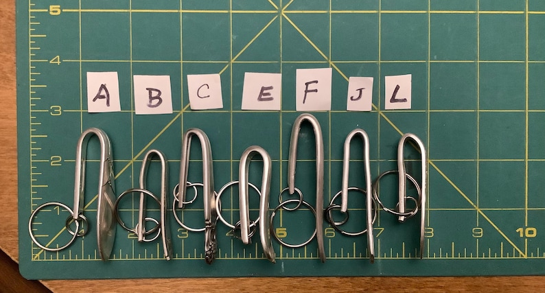 Może przedstawiać: Kolekcja srebrnych metalowych brelok&oacute;w do kluczy o r&oacute;żnych wzorach, w tym brelok w kształcie łyżki. Breloki ułożone są na zielonej macie do cięcia z wzorem kratki i oznaczeniami pomiarowymi.