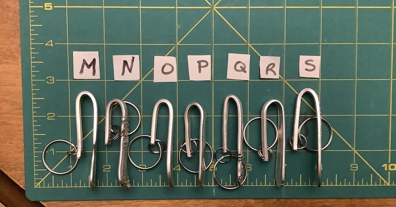 Może przedstawiać: Kolekcja srebrnych brelok&oacute;w do kluczy o kształcie litery U, każdy przymocowany do metalowego k&oacute;łka. Breloki ułożone są na zielonej macie do cięcia, z literami M, N, O, P, Q, R i S napisanymi na małych białych kwadratach powyżej.