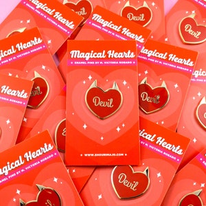 May include: A close-up of several red and gold enamel pins with a heart shape and the word "Devil" in gold lettering. The pins are on a red background with white stars. The text "Magical Hearts" is on the cards with the pins. The text "Enamel Pins by M. Victoria Robado" is also on the cards.