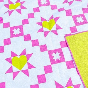 Puede incluir: Un edred&oacute;n hecho a mano con un patr&oacute;n repetido de estrellas rosas con centros de coraz&oacute;n amarillos y acentos de estrellas blancas sobre un fondo blanco. El reverso del edred&oacute;n es de color amarillo verdoso brillante. Un art&iacute;culo de decoraci&oacute;n del hogar acogedor.