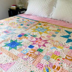 Puede incluir: Un edredón patchwork colorido con diseños de estrellas y espirales. El edredón presenta una variedad de colores pastel, incluyendo rosa, azul, amarillo y verde. Está colocado sobre una cama con almohadas blancas y una colcha rosa.