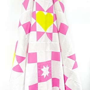 Puede incluir: Un edred&oacute;n hecho a mano con un patr&oacute;n geom&eacute;trico. El edred&oacute;n presenta un fondo de cuadr&iacute;cula blanca con acentos rosa brillante y amarillo. El dise&ntilde;o incluye cuadrados, tri&aacute;ngulos y una forma de coraz&oacute;n central. El edred&oacute;n est&aacute; doblado.