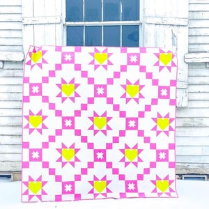 Puede incluir: Un edred&oacute;n hecho a mano con un patr&oacute;n repetitivo de cuadrados y estrellas de color rosa brillante, con corazones amarillos en el centro de las estrellas. El edred&oacute;n se muestra sobre un fondo blanco.