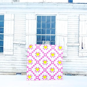Puede incluir: Un edred&oacute;n rosa y blanco con un patr&oacute;n repetido de corazones y estrellas amarillas. El edred&oacute;n se exhibe al aire libre contra un edificio blanco con ventanas y contraventanas, con nieve en el suelo.