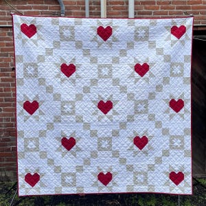 Puede incluir: Un edred&oacute;n blanco con un patr&oacute;n repetido de corazones rojos y formas de estrella beige. El edred&oacute;n tiene un borde rojo y se muestra contra una pared de ladrillo. El edred&oacute;n es probablemente hecho a mano y adecuado para una cama o pared.
