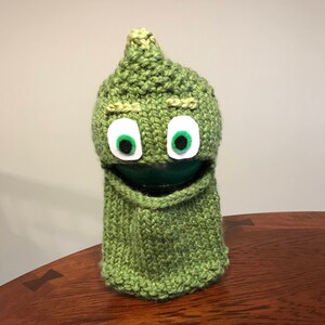 May include: Green knitted hat with a pointed top and a face. The hat has green felt eyes and a brown felt mouth.