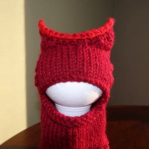 May include: A knitted balaclava in a deep red color, with a contrasting bright red trim around the face opening and the top. The balaclava is displayed on a white styrofoam head, showcasing its shape and design.