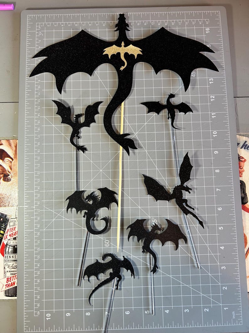 Może przedstawiać: Kolekcja czarnych, brokatowych wycinanek smok&oacute;w na przezroczystych patyczkach, z większym smokiem ze złotym wzorem smoka. Smoki r&oacute;żnią się rozmiarem i pozami, idealne na dekoracje tort&oacute;w.