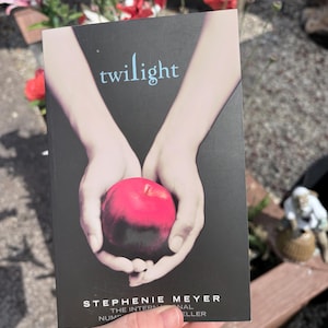 May include: A paperback book titled Twilight by Stephenie Meyer with a black cover featuring a graphic illustration of two pale hands cupped together holding a bright red apple. The title is printed in light blue text at the top of the cover.