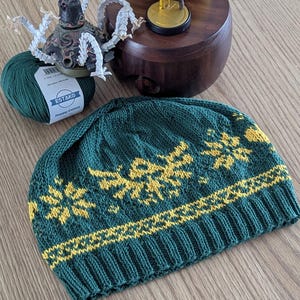 Op de afbeelding: Een groene en gele gebreide muts met een geometrisch patroon. De muts is gemaakt van zacht garen en heeft een comfortabele pasvorm.