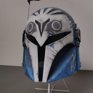 May include: A blue and white Mandalorian helmet with a unique design, featuring a visor and antenna. The helmet has a weathered appearance with black accents and a signature on the side. It is displayed on a clear stand.